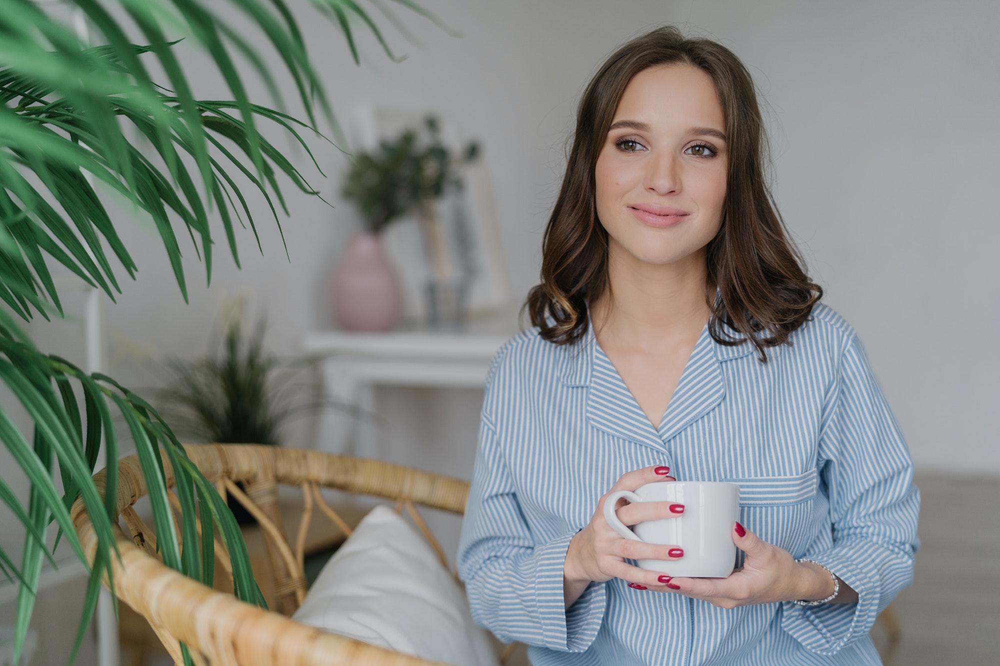 Pleased attractive woman with dark hair, healthy soft skin, drinks coffee in morning